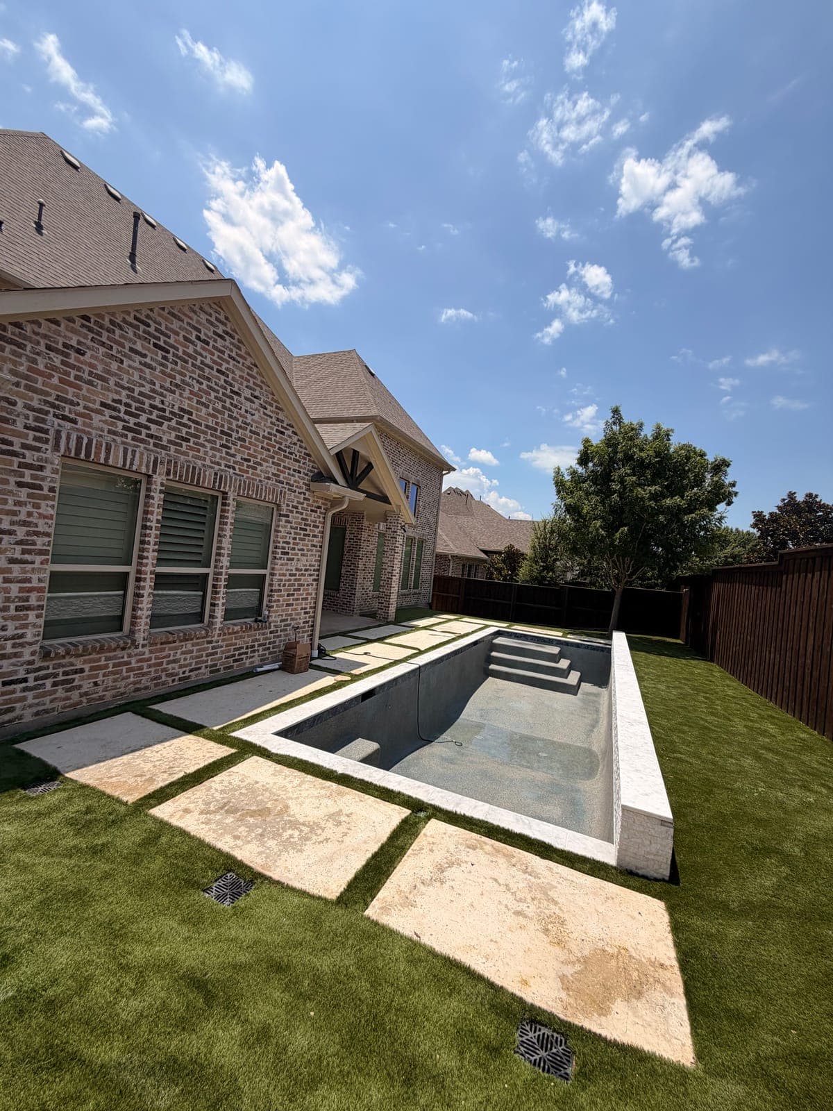 Newly plastered rectangular gunite pool with entry steps and a raised stacked stone wall, set in artificial turf with travertine stepping pads beside a brick home
